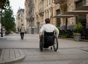 Cilvēks ratiņkrēslā pārvietojas pa Rīgas jūgendstila ielu, kamēr priekšā iet gājējs ar spieķi.