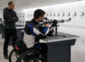 Para šāvējs ratiņkrēslā trenējas ar pneimatisko šauteni iekštelpu šautuvē, trenerim vērojot no malas.