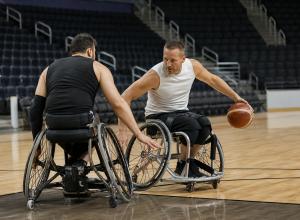 Divi riteņbasketbolisti sporta riteņkrēslos cīnās par bumbu iekštelpu laukumā.