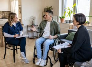Vīrietis riteņkrēslā mājās pārrunā aprūpes plānu ar medicīnas māsu un sociālās aprūpes speciālisti.