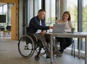 Vīrietis riteņkrēslā un kursu koordinatore pie galda kopīgi pārlūko informāciju klēpjdatorā pieejamā mācību centrā.