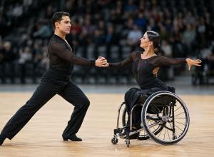Para deju pāris sporta zālē izpilda sinhronu pagriezienu, vienam dejotājam izmantojot riteņkrēslu.