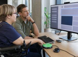 Cilvēks izmanto pielāgotu slēdzi, kamēr kolēģis pārbauda tīmekļa saskarni monitorā.