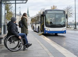 Riteņkrēsla lietotājs un pasažiere pie piekļūstamas pieturas gaida pilsētas autobusu ar izlaistu rampu.