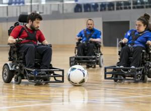 Trīs sportisti elektriskajos riteņkrēslos kontrolē lielu futbola bumbu sporta zālē