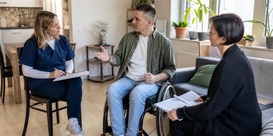 Vīrietis riteņkrēslā mājās pārrunā aprūpes plānu ar medicīnas māsu un sociālās aprūpes speciālisti.