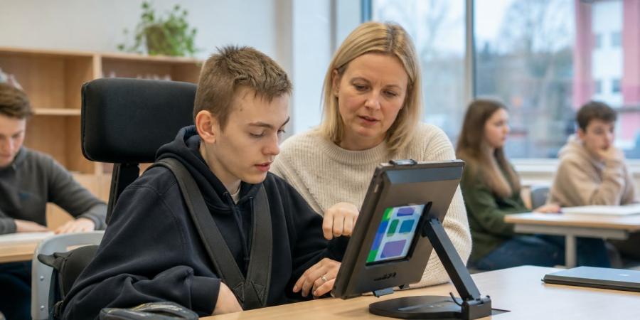 Skolēns klasē kopā ar skolotāju izmanto uz statīva novietotu planšeti ar vizuāliem vadības elementiem.
