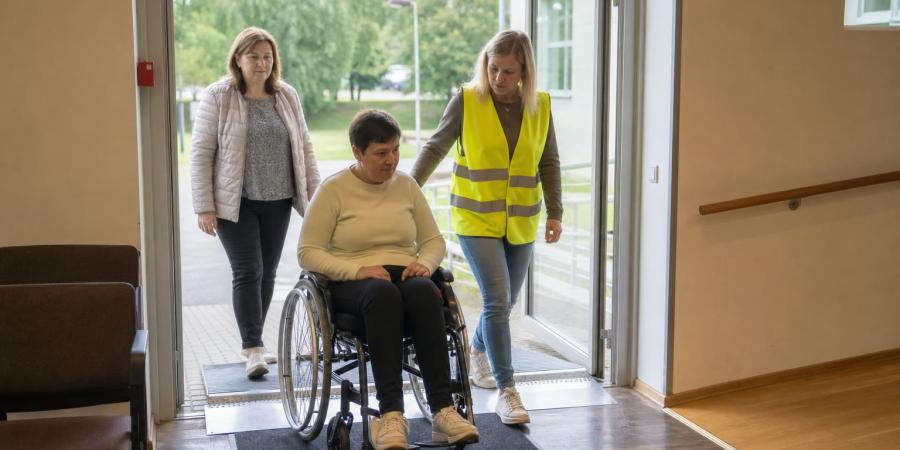 Riteņkrēsla lietotāja ar pavadoni piedalās evakuācijas mācībās, dodoties caur ēkas pieejamo izeju.