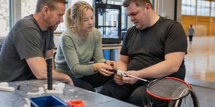 Sportists riteņkrēslā kopā ar inženieri un ergoterapeitu pārbauda personalizētu sporta roktura prototipu.