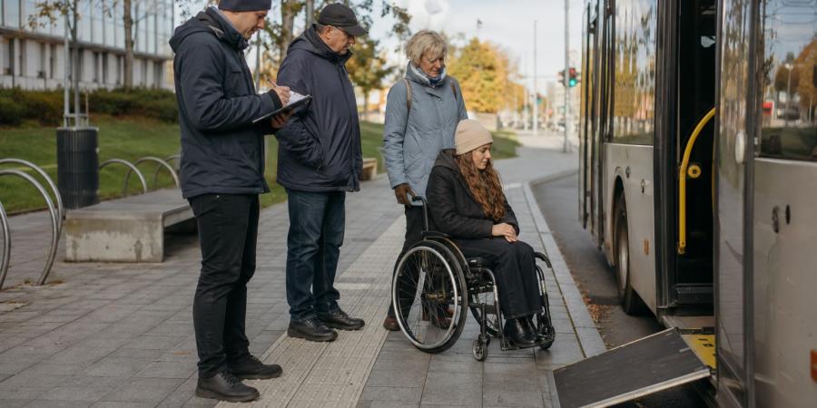 Riteņkrēsla lietotāja un trīs cilvēki pārbauda pieejamību pie pilsētas autobusa pieturas ar izlaistu rampu.