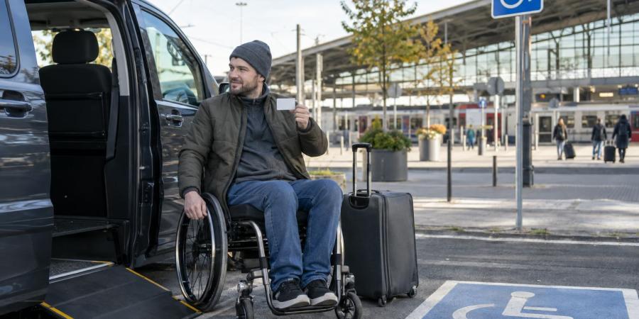 Ceļotājs riteņkrēslā pie pielāgota auto un dzelzceļa stacijas tur rokā karti