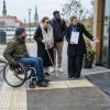 Riteņkrēsla lietotājs un cilvēks ar balto spieķi kopā ar tūrisma darbinieci pārbauda pieejamu ieeju.