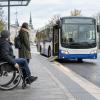 Riteņkrēsla lietotājs un pasažiere pie piekļūstamas pieturas gaida pilsētas autobusu ar izlaistu rampu.