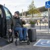 Ceļotājs riteņkrēslā pie pielāgota auto un dzelzceļa stacijas tur rokā karti
