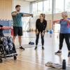 Dažādu paaudžu cilvēki sporta zālē kopīgi vingro ar elastīgajām lentēm