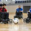 Trīs sportisti elektriskajos riteņkrēslos kontrolē lielu futbola bumbu sporta zālē