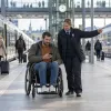 Riteņkrēsla lietotājs dzelzceļa stacijā pārbauda maršrutu, kamēr darbiniece norāda pārsēšanās virzienu