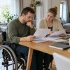 Vīrietis riteņkrēslā un ģimenes locekle pie galda kopīgi pārskata aprūpes izdevumu dokumentus