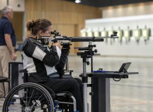 Sportiste riteņkrēslā mērķē ar atbalsta statīvā nostiprinātu šauteni iekštelpu šautuvē
