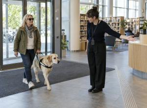 Sieviete ar asistējošo suni ieiet bibliotēkā, kur darbiniece norāda brīvu ceļu