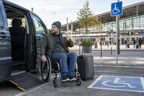 Ceļotājs riteņkrēslā pie pielāgota auto un dzelzceļa stacijas tur rokā karti