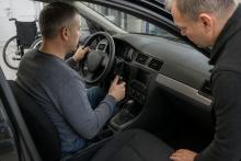 Cilvēks riteņkrēslā kopā ar tehniķi darbnīcā apskata automobilī uzstādītu rokas vadības ierīci.