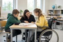 Skolniece ratiņkrēslā pie galda kopā ar skolotāju un klasesbiedru pārrunā mācību uzdevumu gaišā klasē.