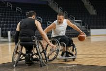 Divi riteņbasketbolisti sporta riteņkrēslos cīnās par bumbu iekštelpu laukumā.