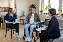 Vīrietis riteņkrēslā mājās pārrunā aprūpes plānu ar medicīnas māsu un sociālās aprūpes speciālisti.