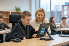 Skolēns klasē kopā ar skolotāju izmanto uz statīva novietotu planšeti ar vizuāliem vadības elementiem.