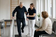 Fizioterapeite rehabilitācijas zālē palīdz vecākam vīrietim vingrināt līdzsvaru, kamēr tuviniece vēro nodarbību.