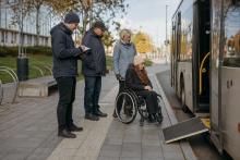 Riteņkrēsla lietotāja un trīs cilvēki pārbauda pieejamību pie pilsētas autobusa pieturas ar izlaistu rampu.