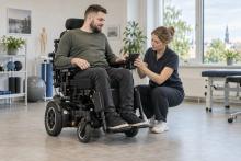 Elektriskā riteņkrēsla lietotājs kopā ar ergoterapeiti regulē vadības ierīci rehabilitācijas telpā.