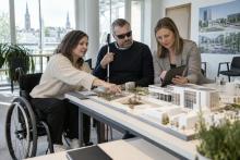 Riteņkrēsla lietotāja, cilvēks ar balto spieķi un speciāliste kopā izvērtē ēkas maketu un planšeti.