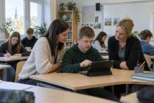 Skolēns klasē lieto planšetdatoru, kamēr skolotāja un atbalsta speciāliste palīdz izvēlēties mācību darbību.