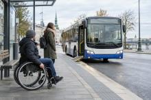 Riteņkrēsla lietotājs un pasažiere pie piekļūstamas pieturas gaida pilsētas autobusu ar izlaistu rampu.