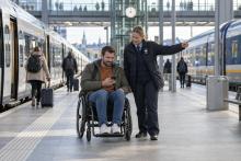 Riteņkrēsla lietotājs dzelzceļa stacijā pārbauda maršrutu, kamēr darbiniece norāda pārsēšanās virzienu