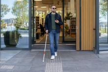 Neredzīgs cilvēks ar balto spieķi un tālruni pārvietojas pa taktilo vadlīniju pie bibliotēkas ieejas