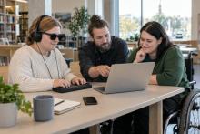 Trīs cilvēki bibliotēkā kopīgi pārbauda piekļūstamu digitālo pakalpojumu datorā un taktilā ierīcē