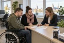Riteņkrēsla lietotājs un asistente kopā ar sociālo darbinieci pārskata pakalpojuma grafiku