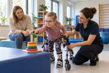 Meitene ar staigāšanas palīgierīci rehabilitācijas nodarbībā sniedzas pēc krāsaina riņķa