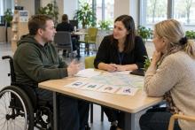 Vīrietis riteņkrēslā ar atbalsta personu pārrunā pakalpojumus pie sociālās darbinieces