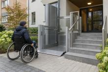 Cilvēks riteņkrēslā pie daudzdzīvokļu mājas ieejas ar platformas pacēlāju un kāpnēm