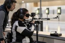 Sportiste ar aizsegtām acīm un austiņām mērķē ar pneimatisko šauteni trenera klātbūtnē