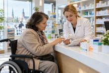 Sieviete riteņkrēslā kopā ar farmaceiti pie letes pārskata zāļu iepakojumu un lietošanas plānu