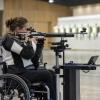 Sportiste riteņkrēslā mērķē ar atbalsta statīvā nostiprinātu šauteni iekštelpu šautuvē