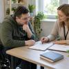 Vīrietis riteņkrēslā kopā ar speciālisti pārskata rehabilitācijas plānu pie galda