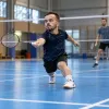Maza auguma sportists izklupienā atsit volānu badmintona laukumā, treniņu partnerim stāvot fonā