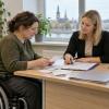 Sieviete riteņkrēslā kopā ar sociālo darbinieci pārskata čekus un pabalsta dokumentus