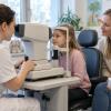 Acu aprūpes speciāliste pārbauda meitenes redzi, kamēr mamma sēž līdzās